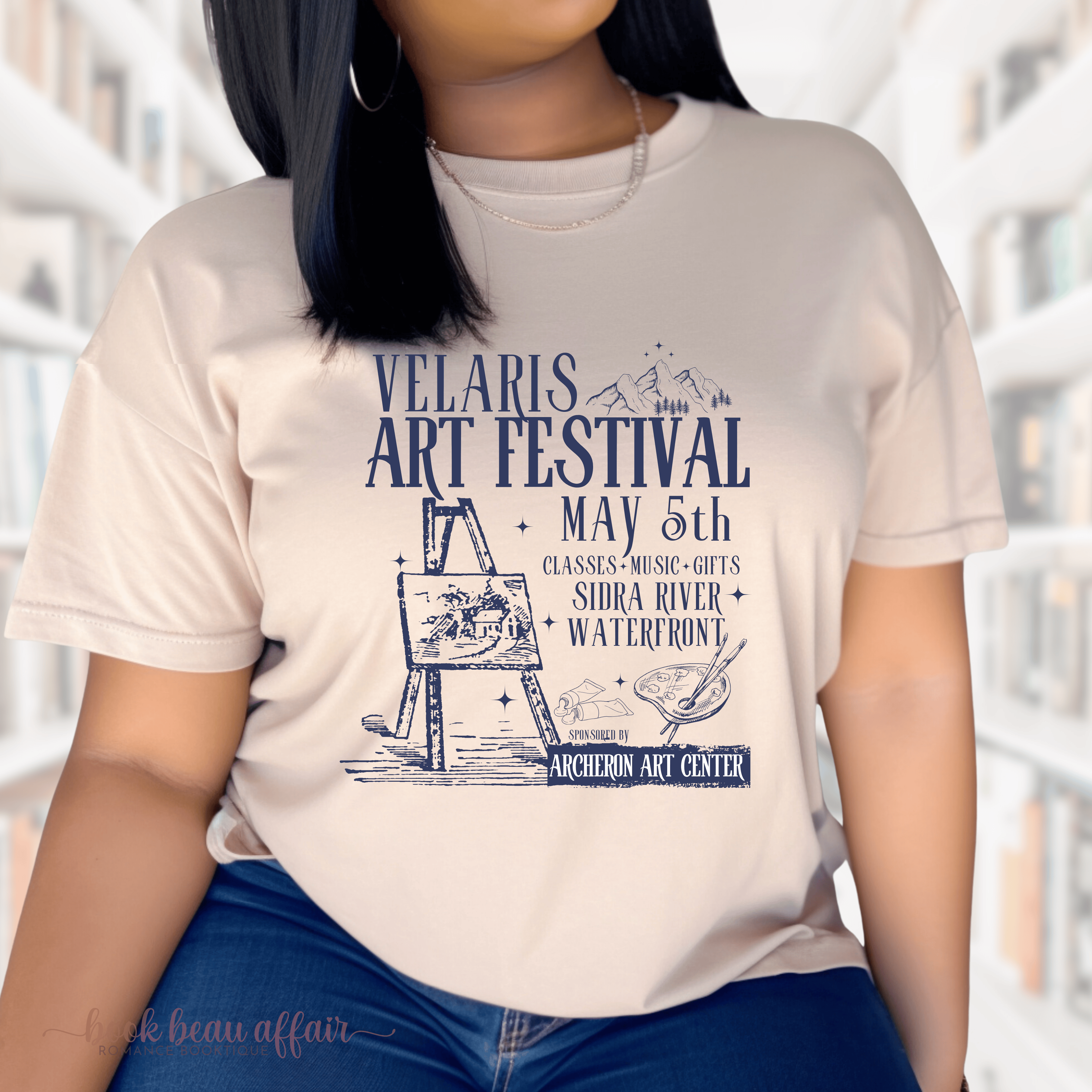 A person wearing a sand t-shirt with 'VELARIS ART FESTIVAL' and other details printed on it, standing in front of a bookshelf.