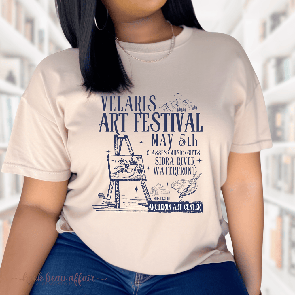 A person wearing a sand t-shirt with 'VELARIS ART FESTIVAL' and other details printed on it, standing in front of a bookshelf.