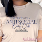 Close up of A woman wearing a light sand colored graphic t-shirt with navy blue text that reads 'MEMBERS ONLY ANTI SOCIAL Book Club' and an illustration of an open book. 