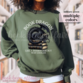 military green sweatshirt with a dragon laying on a stack of books and text that says book dragon, like a bookworm but way cooler