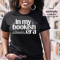 Person wearing a black t-shirt with 'In my bookish era' text and book graphic, standing in a library.