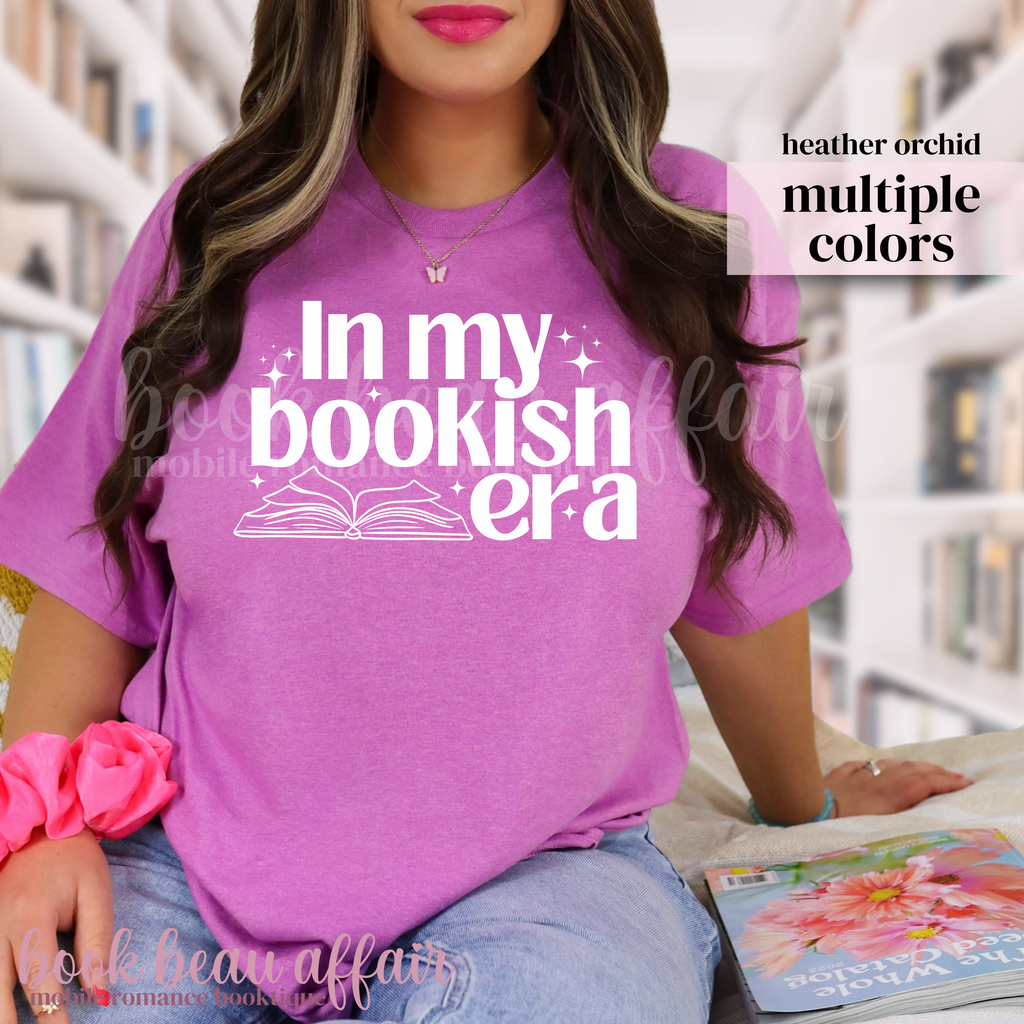 Person wearing a pink t-shirt with 'In my bookish era' text, sitting in a library.