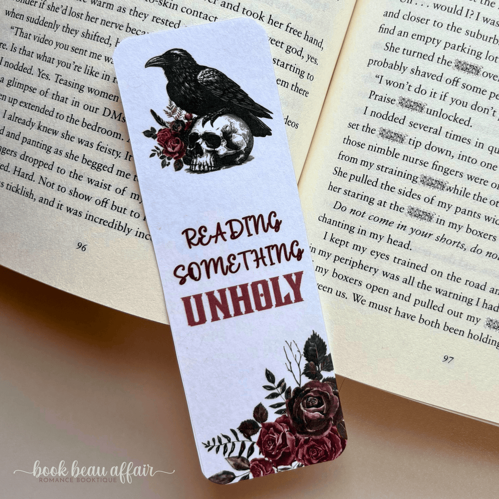 Bookmark with a raven and skull design on an open book that says reading something unholy
