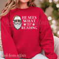 Red sweatshirt with Santa Claus graphic and text 'He sees what you've been reading' on a blurred background.