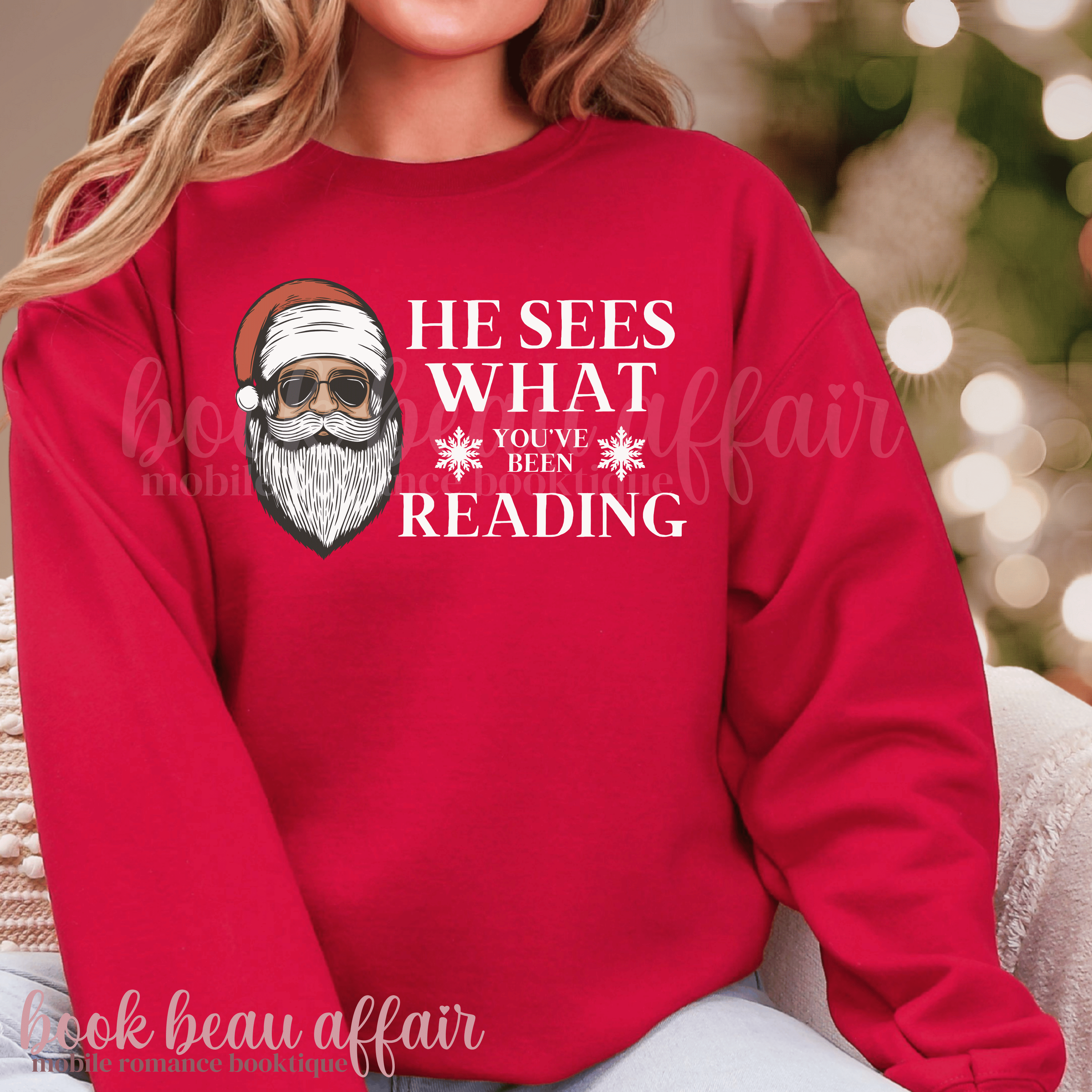 Red sweatshirt with Santa Claus graphic and text 'He sees what you've been reading' on a blurred background.