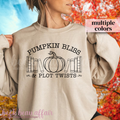 Person wearing a beige sweatshirt with 'Pumpkin Bliss & Plot Twists' text and pumpkin graphic, with autumn leaves in the background.