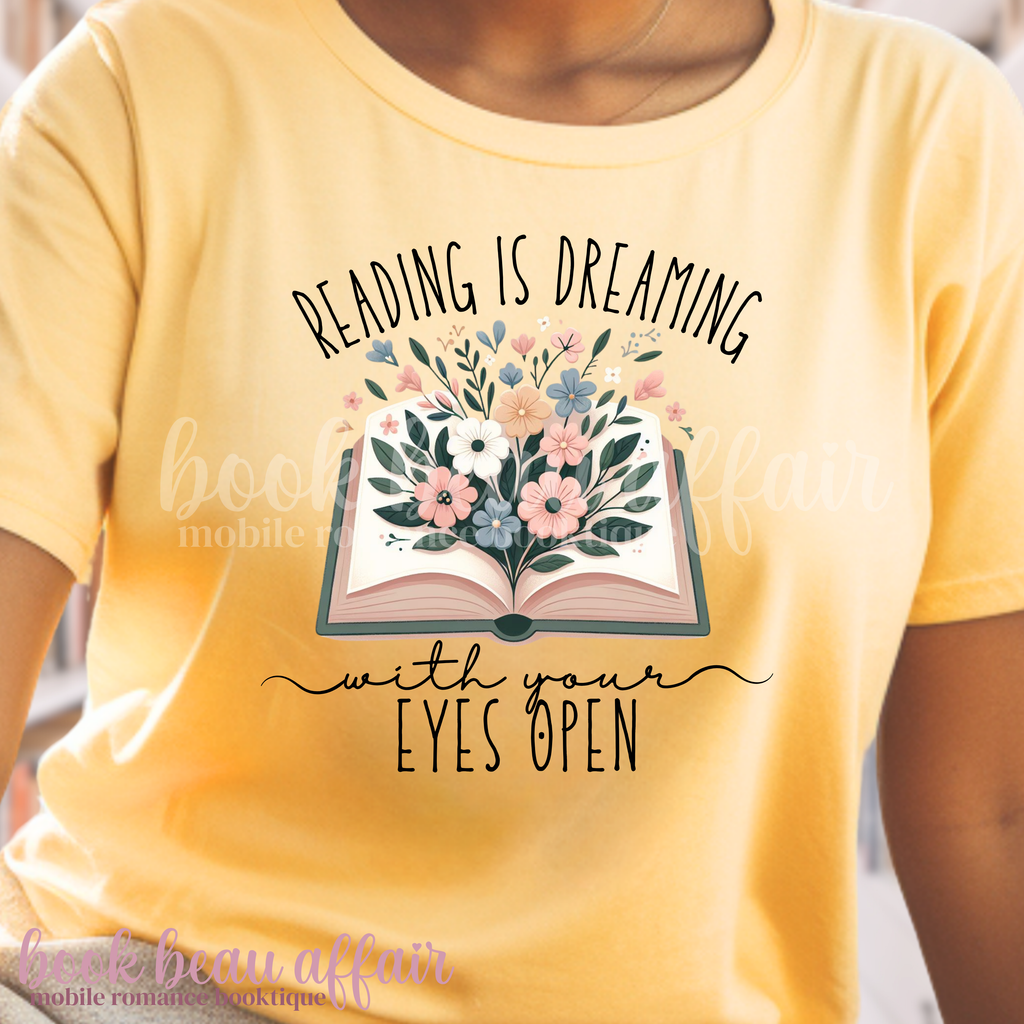 Yellow t-shirt with floral book graphic and text 'Reading is dreaming with your eyes open' on a blurred background.