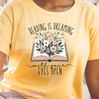 Yellow t-shirt with floral book graphic and text 'Reading is dreaming with your eyes open' on a blurred background.