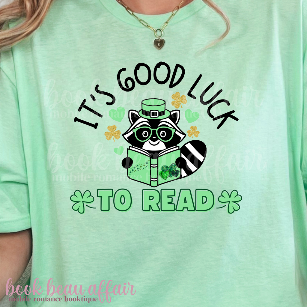 St. Patricks Day Themed Shirt. Illustration of a raccoon holding a green book with text that says "It's Good Luck to Read"