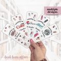 Hand holding multiple bookmarks with various diet soda brand logos and text, with a blurred indoor background.