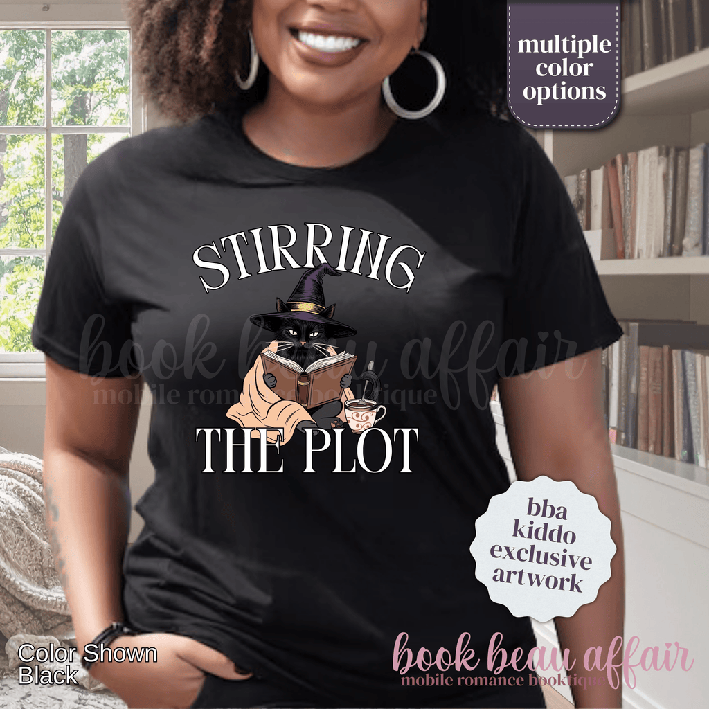 A woman wearing a black graphic t-shirt with the text 'STIRRING THE PLOT' and an illustration of a cat in a witch hat reading a book. 