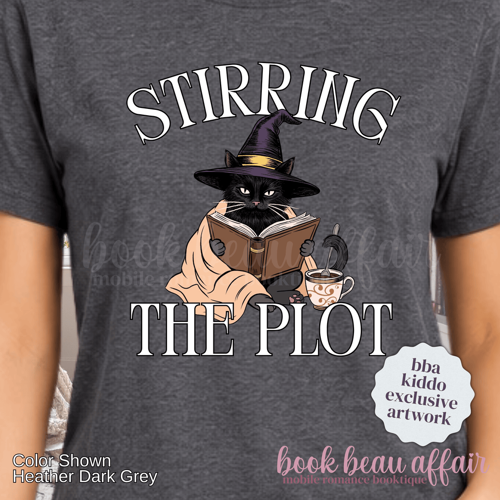 close up of a dark gray graphic t-shirt with the text 'STIRRING THE PLOT' and an illustration of a cat in a witch hat reading a book. 