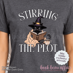 close up of a dark gray graphic t-shirt with the text 'STIRRING THE PLOT' and an illustration of a cat in a witch hat reading a book. 