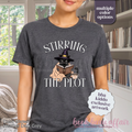 A woman wearing a dark gray graphic t-shirt with the text 'STIRRING THE PLOT' and an illustration of a cat in a witch hat reading a book. 