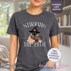 A woman wearing a dark gray graphic t-shirt with the text 'STIRRING THE PLOT' and an illustration of a cat in a witch hat reading a book. 