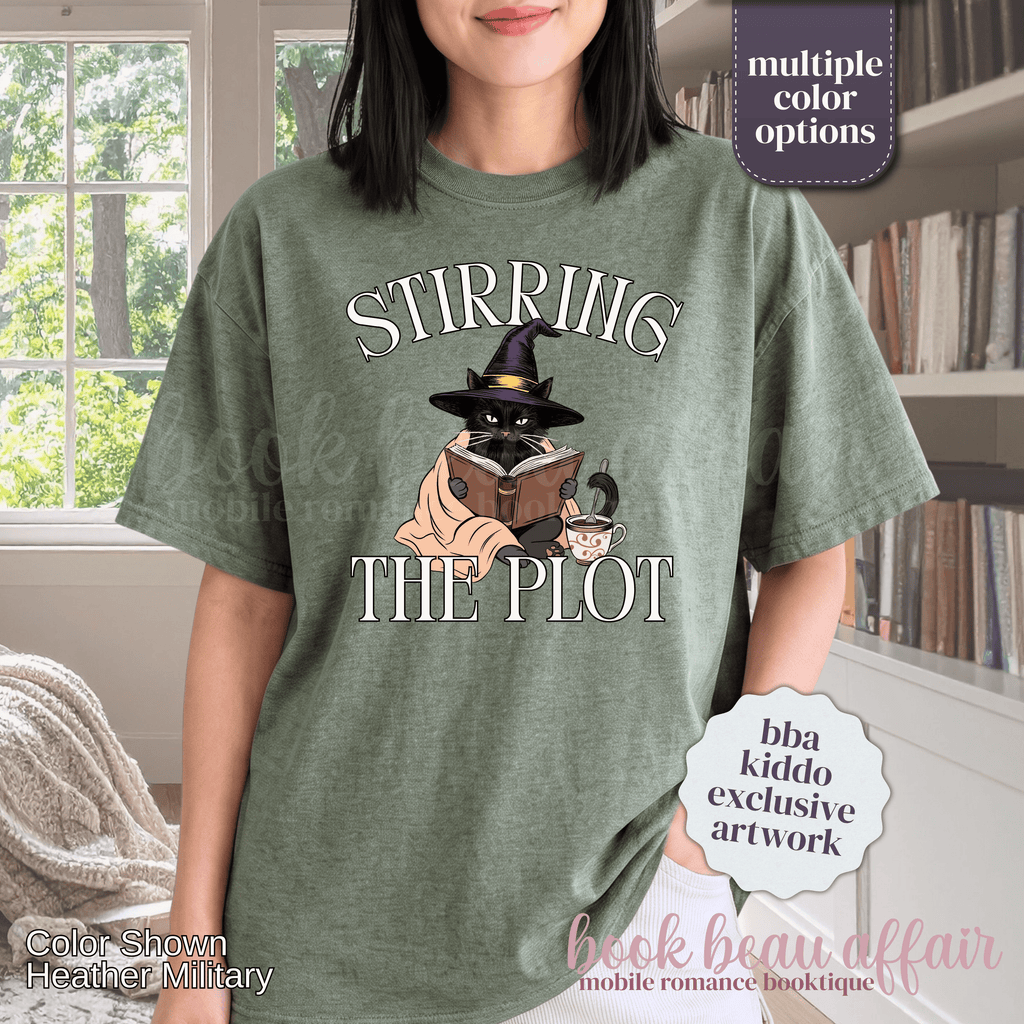 A woman wearing a heather green graphic t-shirt with the text 'STIRRING THE PLOT' and an illustration of a cat in a witch hat reading a book. 