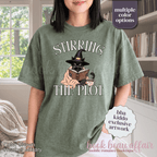 A woman wearing a heather green graphic t-shirt with the text 'STIRRING THE PLOT' and an illustration of a cat in a witch hat reading a book. 