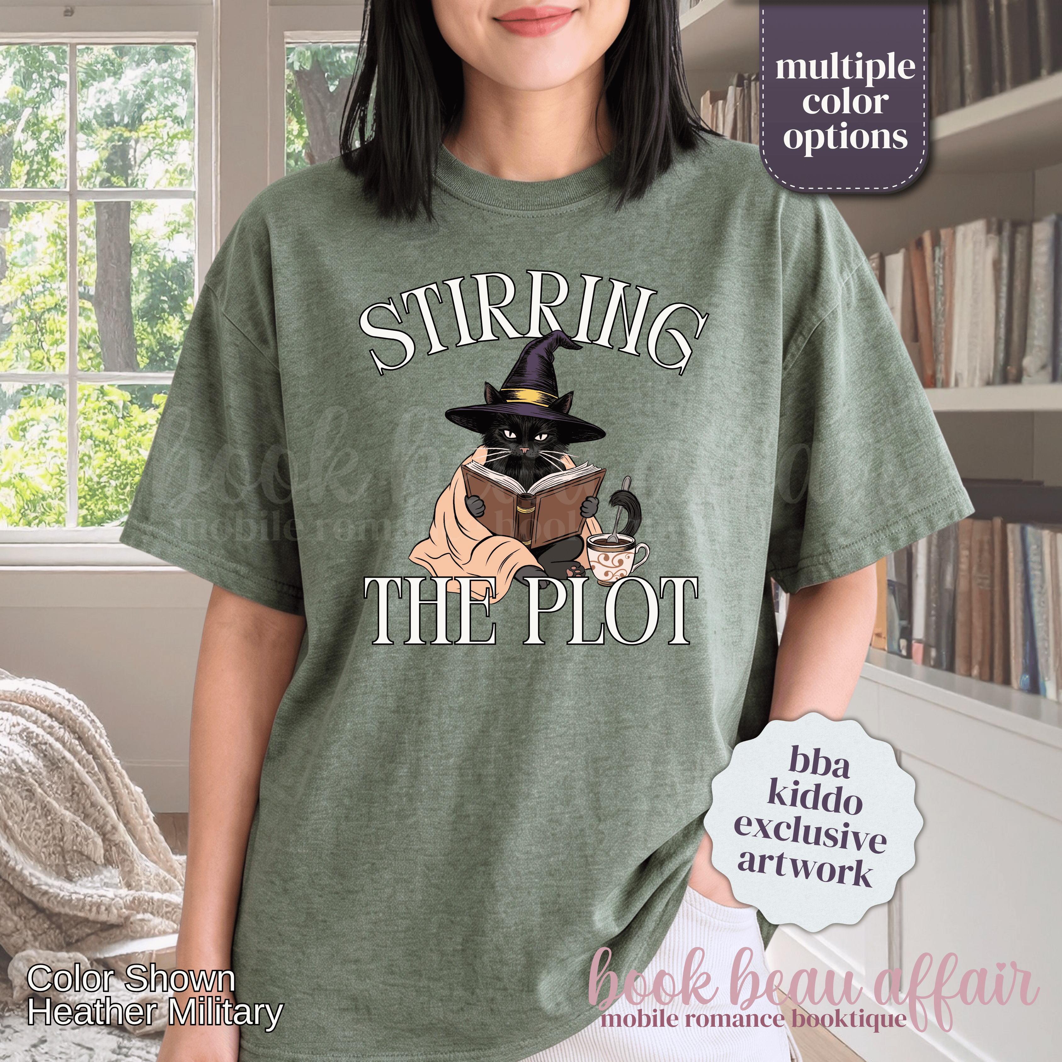 A woman wearing a heather green graphic t-shirt with the text 'STIRRING THE PLOT' and an illustration of a cat in a witch hat reading a book. 