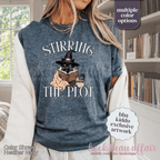 A woman wearing a heather navy graphic t-shirt with the text 'STIRRING THE PLOT' and an illustration of a cat in a witch hat reading a book. 