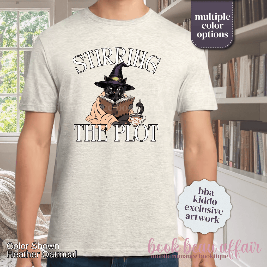 A man wearing a heather oatmeal graphic t-shirt with the text 'STIRRING THE PLOT' and an illustration of a cat in a witch hat reading a book. 