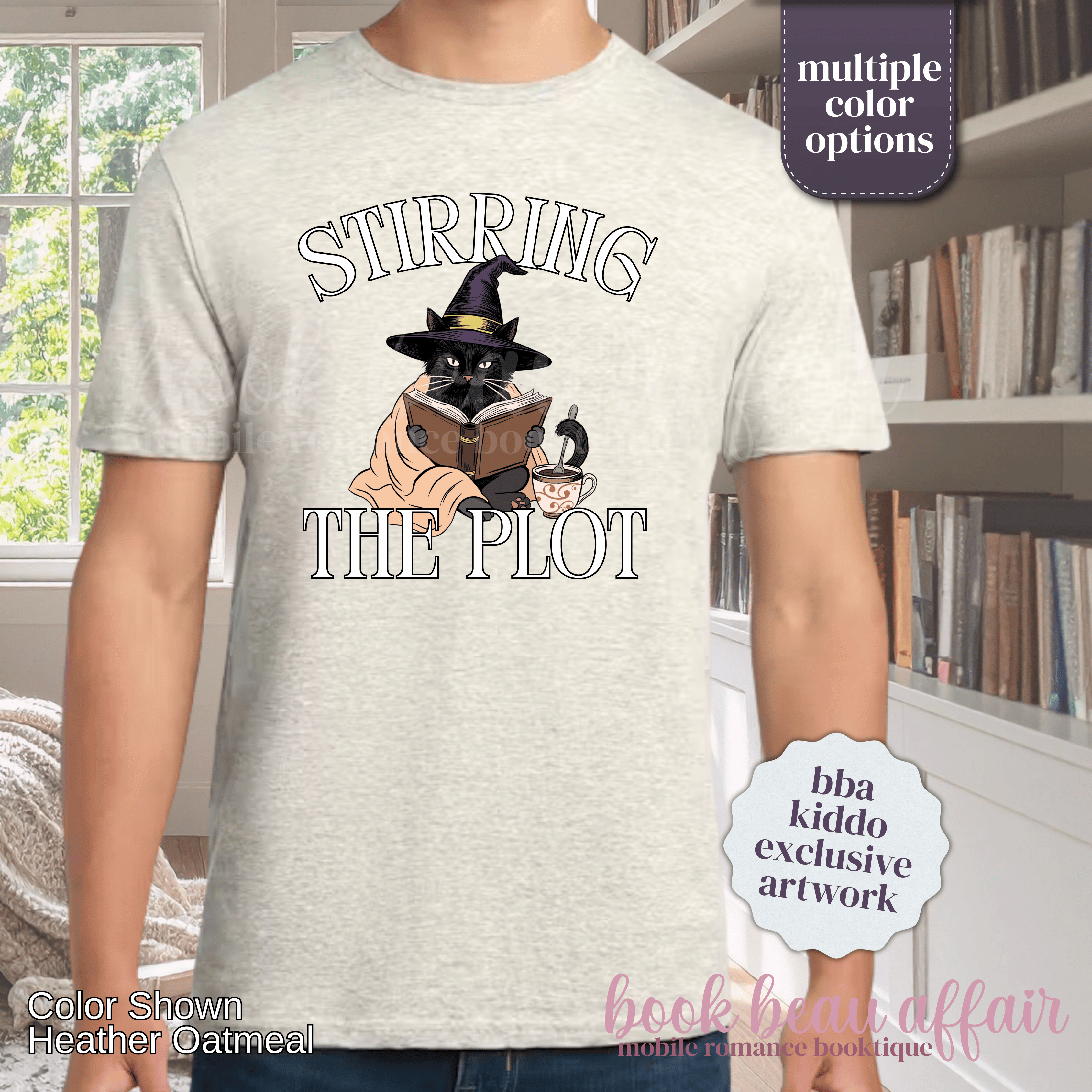 A man wearing a heather oatmeal graphic t-shirt with the text 'STIRRING THE PLOT' and an illustration of a cat in a witch hat reading a book. 