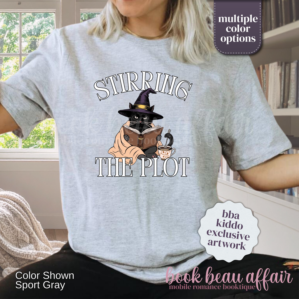 A woman wearing a light gray graphic t-shirt with the text 'STIRRING THE PLOT' and an illustration of a cat in a witch hat reading a book. 