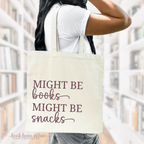 A person holding a beige cotton tote bag with the text 'MIGHT BE BOOKS. MIGHT BE SNACKS.