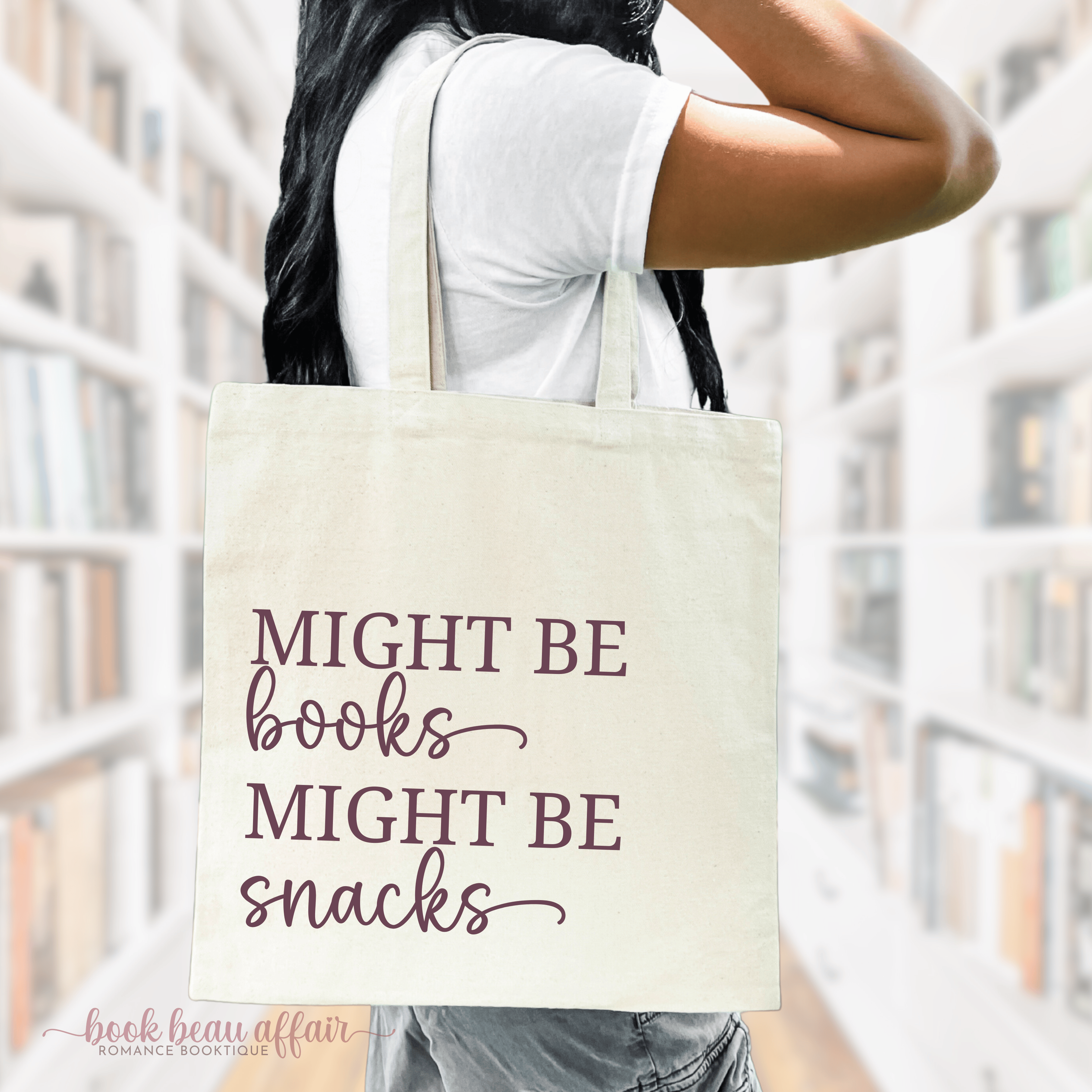 A person holding a beige cotton tote bag with the text 'MIGHT BE BOOKS. MIGHT BE SNACKS.